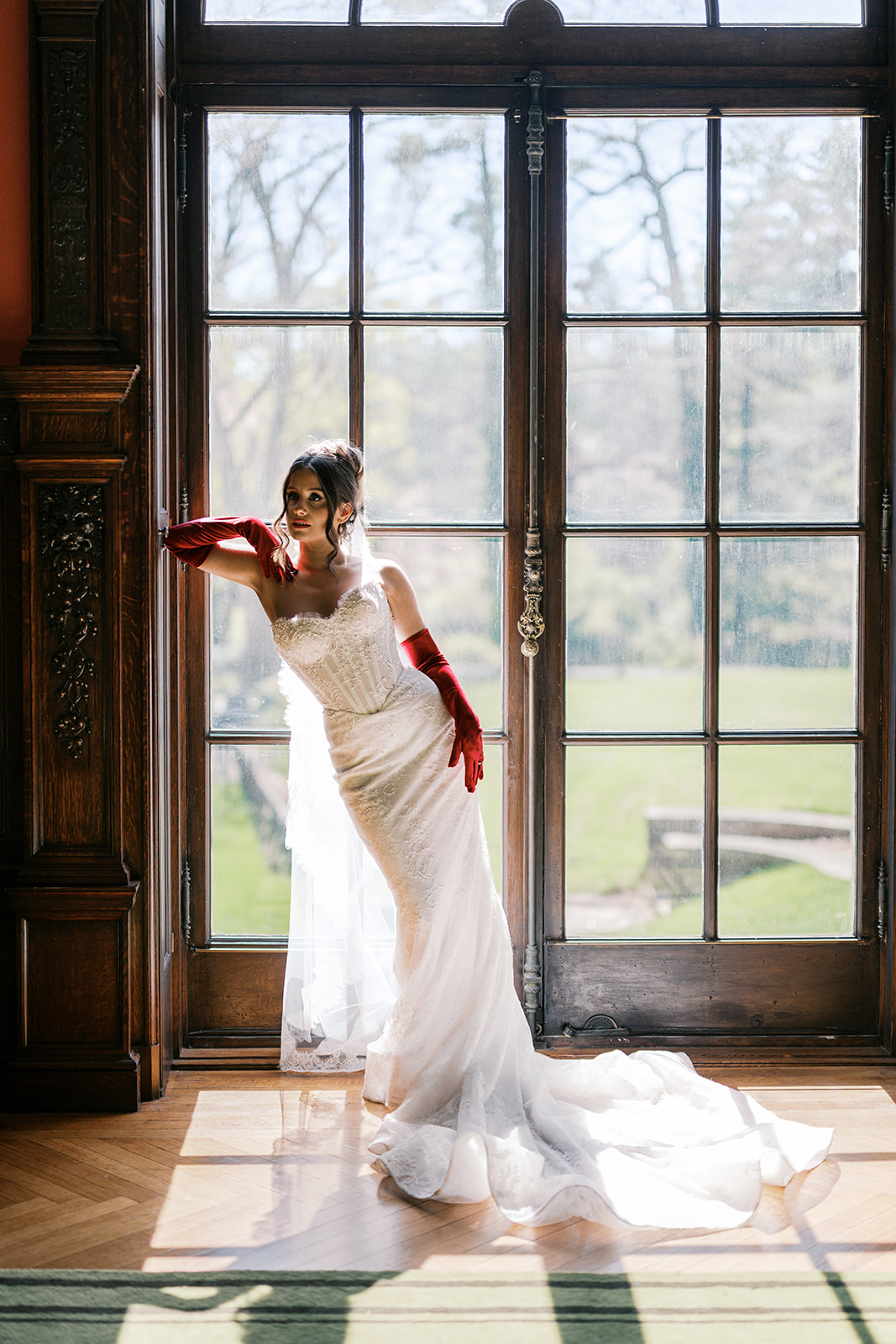 Bride wearing a basque waist wedding dress and red elbow length gloves leans up against a door frame in front of wooden french doors at Elkins Estate