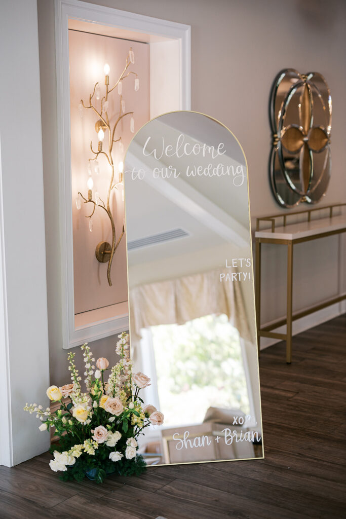 Arched mirror with the words "Welcome to Our Wedding", "Let's Party!" and "xoxo Shan + Brian" sits next to a pastel pink and yellow floral arrangement | Lindsey Ford Photography