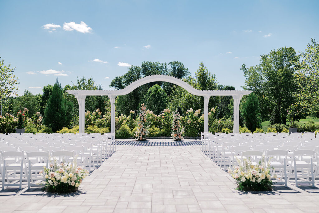 Two pastel pink, orange, and white floral column installs sit under a white pergola on a stone patio facing rows of white ceremony chairs | Lindsey Ford Photography at The Gardens at Applecross