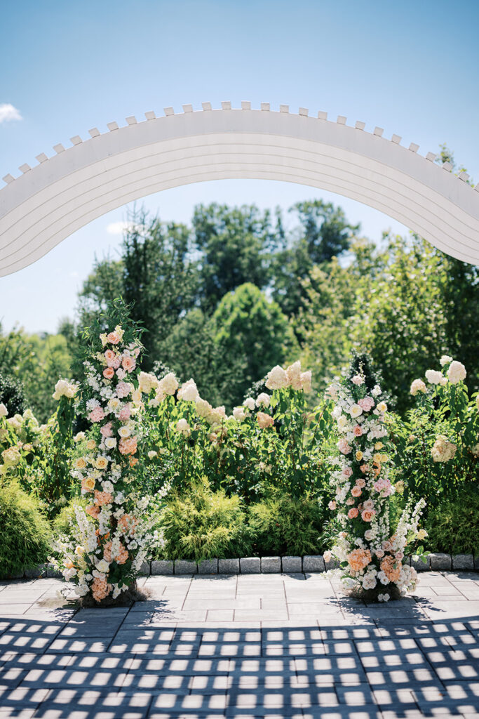 Two pastel pink, orange, and white floral column installs sit under a white pergola on a stone patio at The Gardens at Applecross | Lindsey Ford Photography