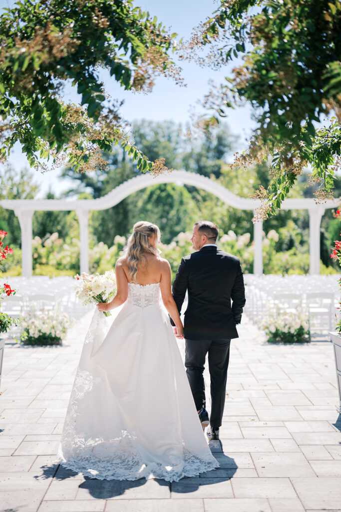 Bride and groom hold hands and walk toward the front of their outdoor ceremony space which is framed by an arched white pergola | Lindsey Ford Photography
