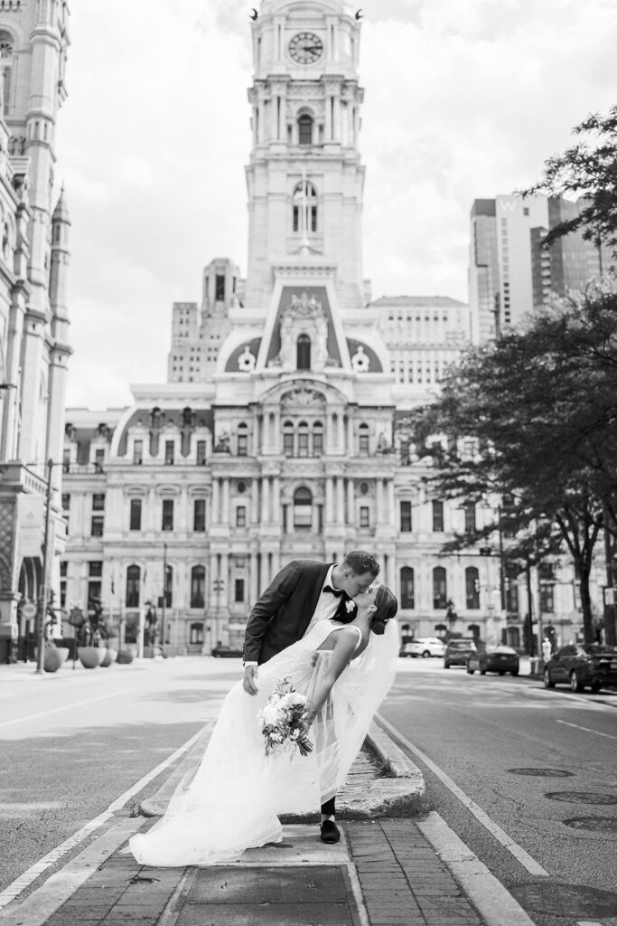 Black and white photo of groom dipping bride in front of the Philadelphia City Hall building during their wedding portraits