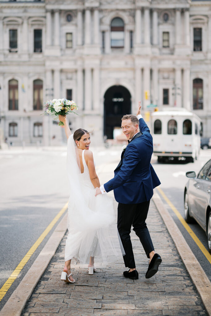Bride and groom hold hands and walk in the middle of the road in front of Philadelphia City Hall building | Lindsey Ford Photography