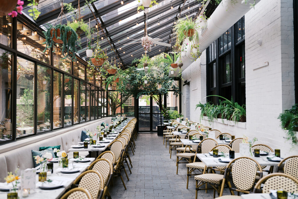Outdoor patio at Osteria in Philly is filled with two rows of wedding reception tables set with plates underneath hanging greenery and market lights