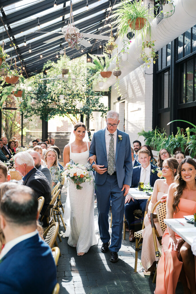 Father walks bride down the aisle for her wedding ceremony surrounded by wedding guests at tables inside the glass patio at Osteria in Philly