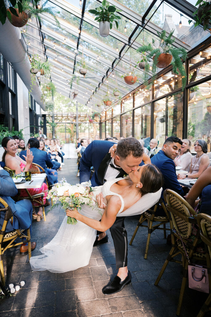 Groom dips bride and kisses her while guests clap and cheer underneath hanging greenery and market lights at the Osteria restaurant patio in Philly