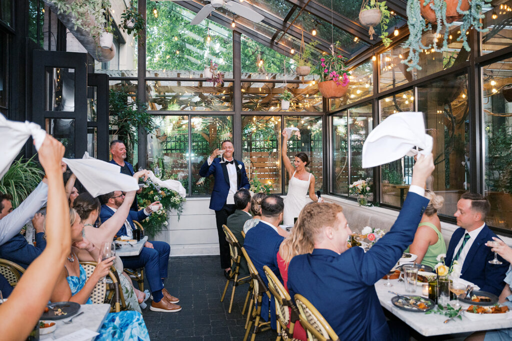 Wedding guests wave white napkins in the air as bride and groom stand up and toast the room