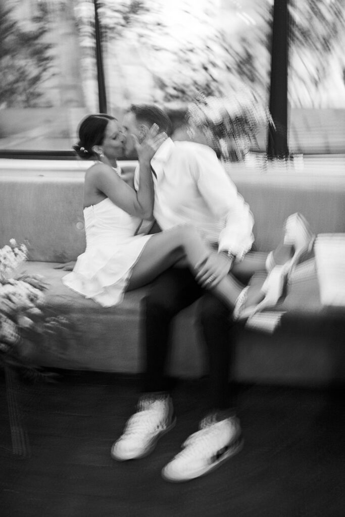 Bride in white mini dress and platform white heels sits on grooms lap as they kiss | Black and white motion blur wedding photography by Lindsey Ford