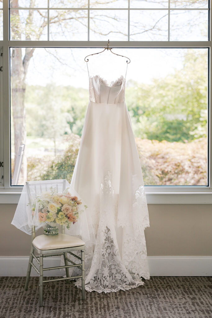 Strapless wedding dress hangs in front of a wide window sitting next to a chair holding bride's pastel bouquet and veil