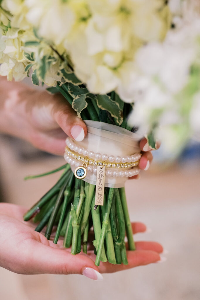 Close up photo of bouquet stems wrapped in a cream colored ribbon, a string of pearls and gems, with a small blue charm