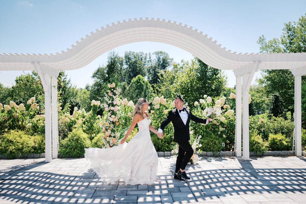 Bride and groom hold hands and run underneath a white arched pergola wedding ceremony backdrop at The Gardens at Applecross