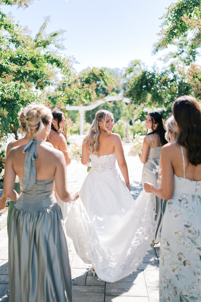 Bride walks with six bridesmaids in sage green dresses as they hold the train of her lace wedding dress