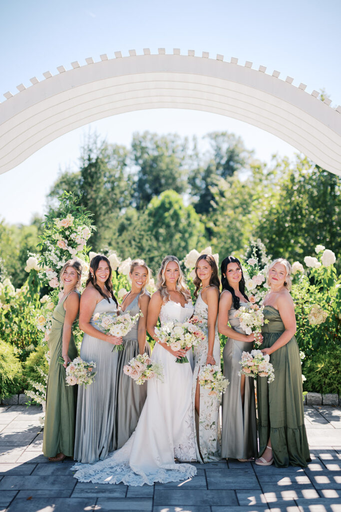 Bride stands with six bridesmaids in sage green dresses underneath a white arched pergola wedding ceremony backdrop at The Gardens at Applecross