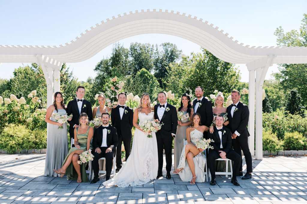 Bride and groom stand surrounded by their wedding party underneath a white arched pergola wedding ceremony backdrop at The Gardens at Applecross