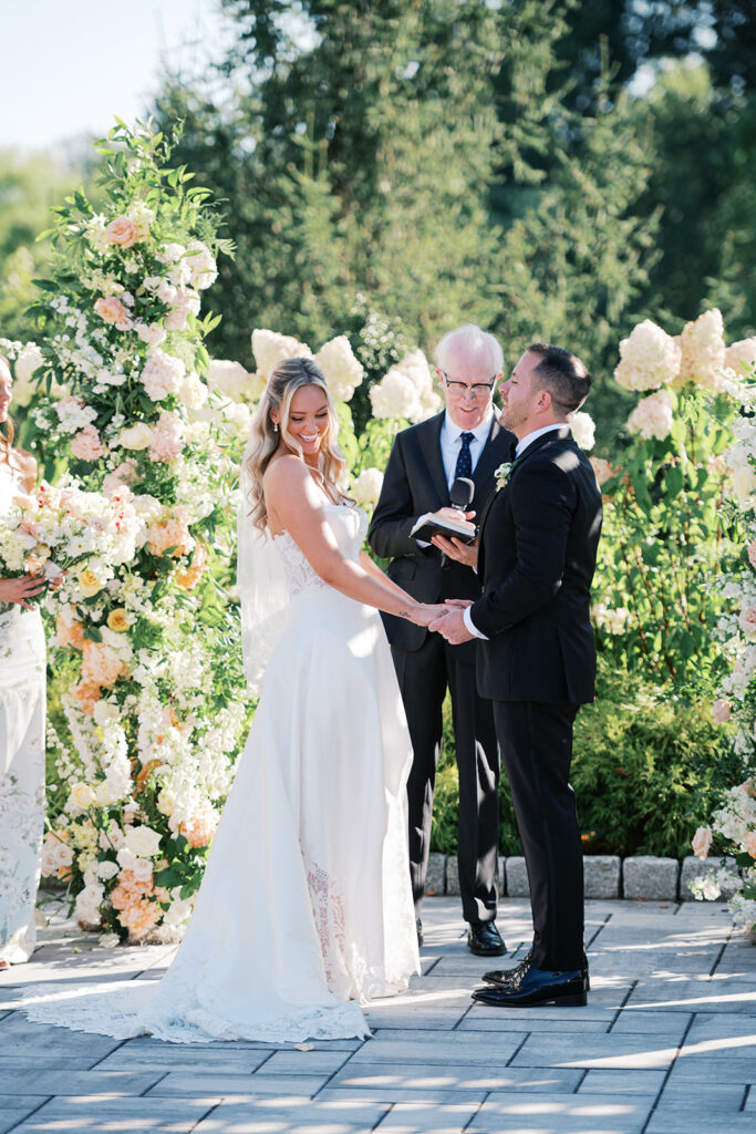 Bride and groom stand with officiant in front of a large hedge of white hydrangeas at the Gardens at Applecross