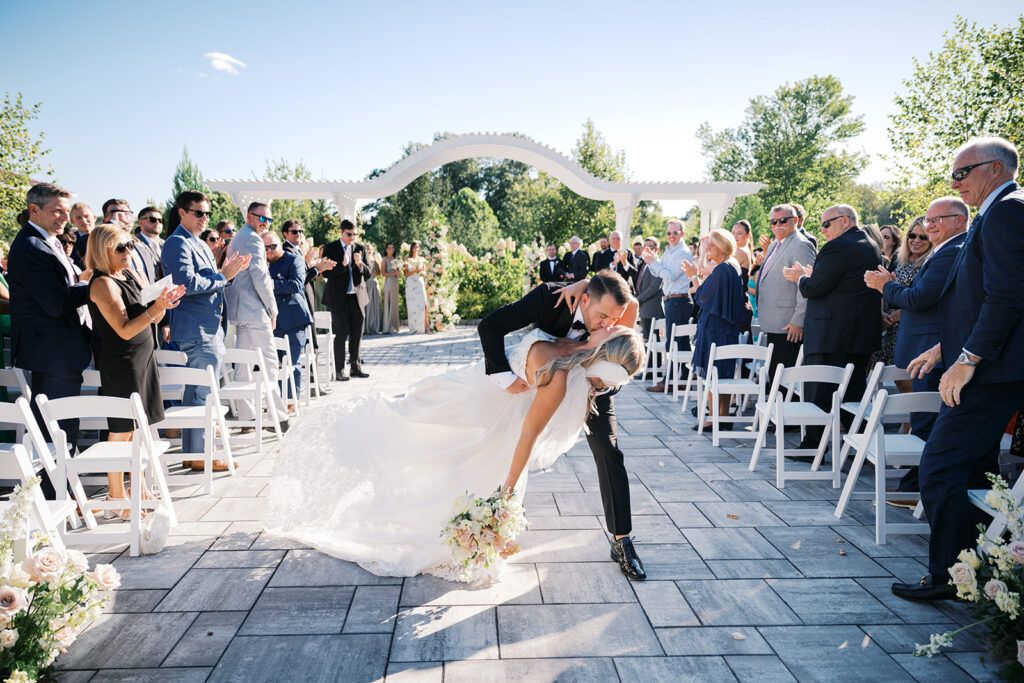 Groom dips bride in the aisle of their wedding ceremony in front of a white arched pergola while guests watch and clap at The Gardens at Applecross