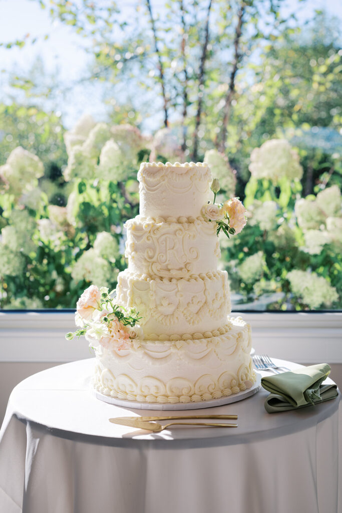 Four tier white wedding cake decorated with intricate piped icing details sits in front of a window