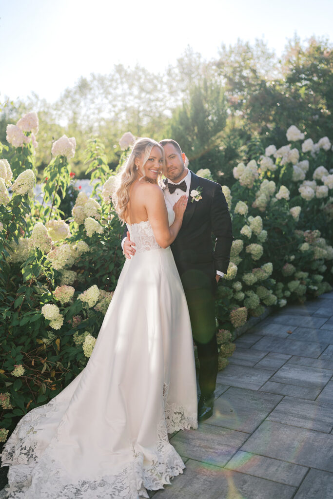 Bride and groom stand together and pose in front of a hedge of white hydrangeas at The Gardens at Applecross