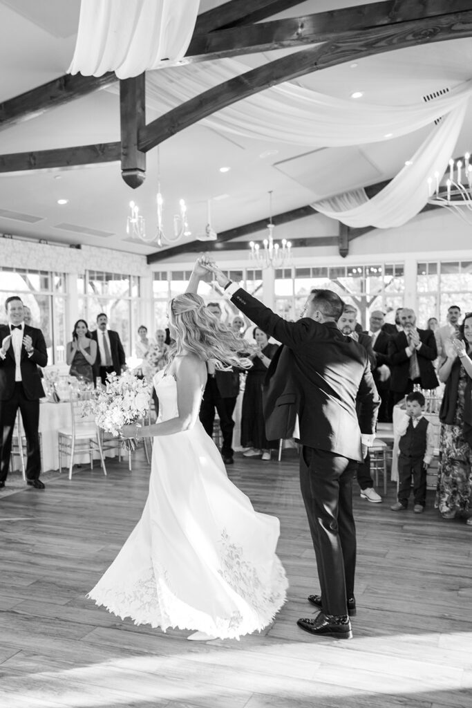 Groom twirls bride on the dance floor inside the reception hall at The Gardens at Applecross while guests watch and clap