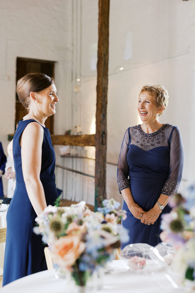 Two women in navy gowns talk and laugh surrounded by floral arrangements before a wedding | Lindsey Ford Photography