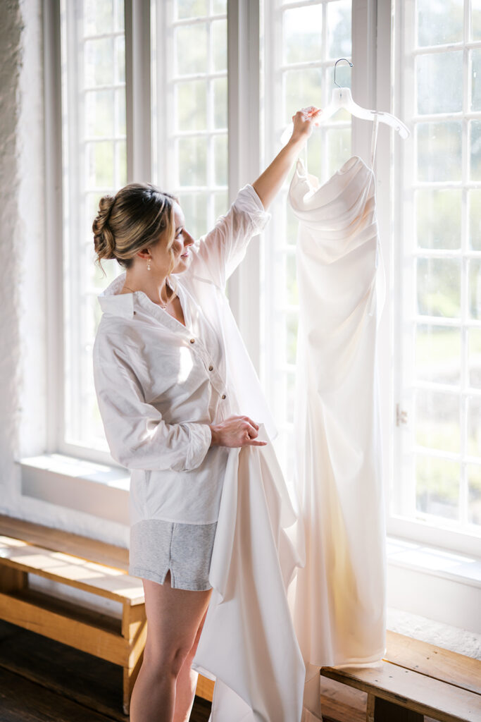 Bride holds up her wedding dress on a hanger in front of a window at Bartram's Garden wedding venue in Philadelphia | Lindsey Ford Photography