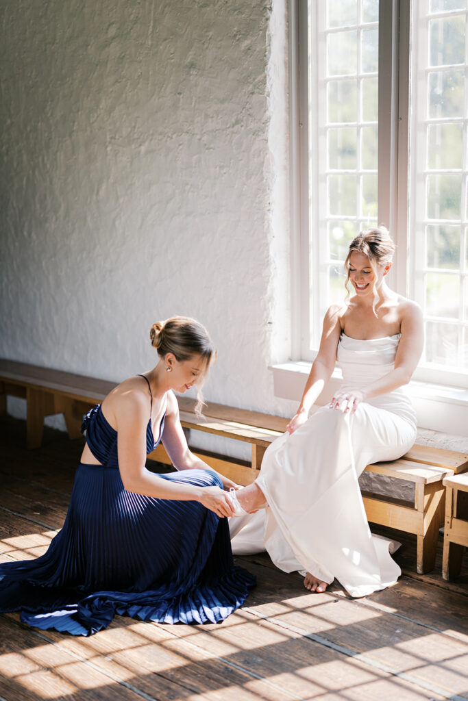 Girl in navy gown helps bride put on her shoe while bride sits on a wooden bench in front of a window at Bartram's Garden in Philadelphia | Lindsey Ford Photography