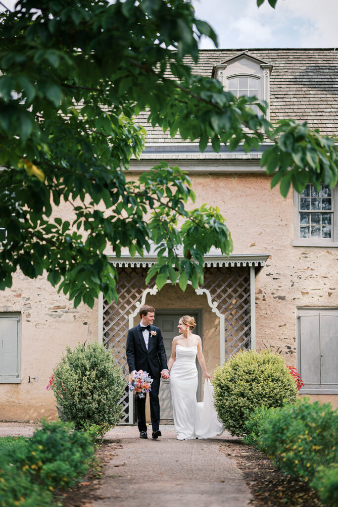 Bride and groom walk hand in hand in front of a historic stone building at the Bartram's Garden wedding venue in Philadelphia