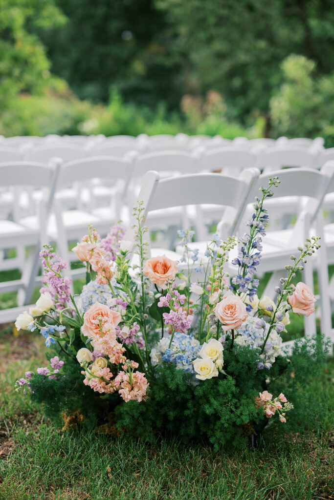 Pastel colored floral arrangement sits on the ground behind rows of white folding ceremony chairs for a wedding at Bartram's Garden