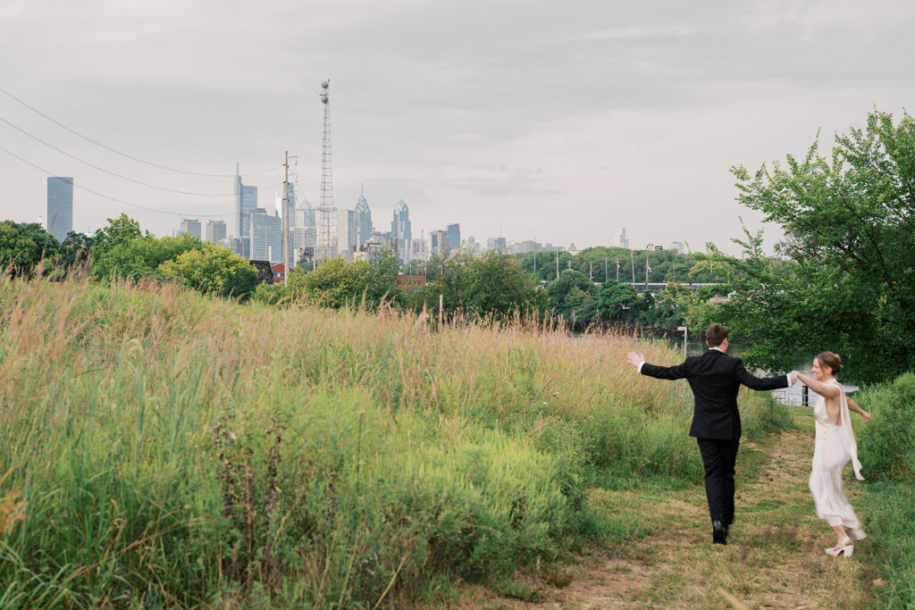 Bride and groom run hand in hand in a grassy field in front of the Philly skyline during their wedding portraits at Bartram's Garden by Lindsey Ford Photography