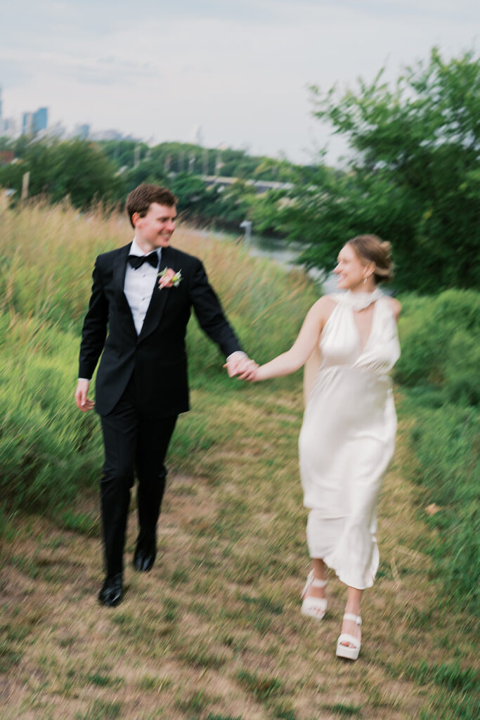 Bride and groom run hand in hand in a grassy field during their wedding portraits at Bartram's Garden by Lindsey Ford Photography in Philadelphia