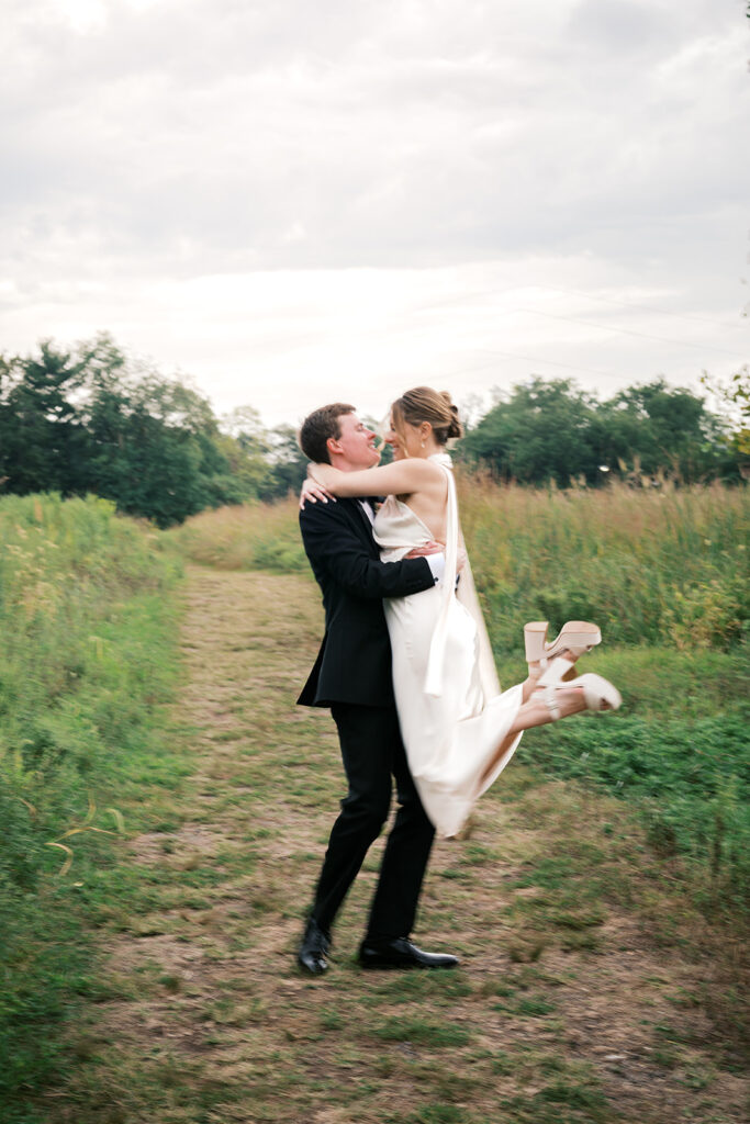 Groom lifts bride in the middle of a grassy field during their wedding portraits at Bartram's Garden by Lindsey Ford Photography in Philadelphia