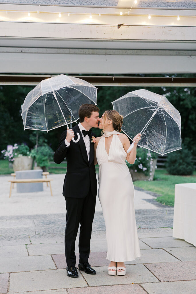 Bride and groom kiss while holding clear umbrellas under a reception tent at Bartram's Garden in Philadelphia