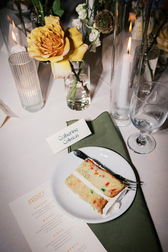 A plate with a slice of funfetti cake sits on a green napkin on a wedding reception table surrounded by pillar candles and flowers at Bartram's Gardens