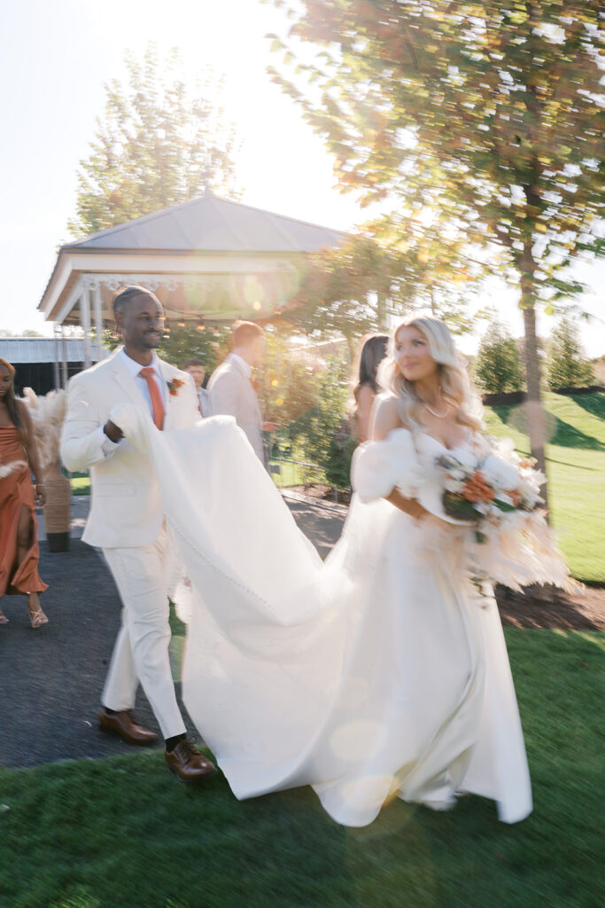 Groom holds the train of brides dress as they walk. Light flares from the setting sun cast a warm glow
