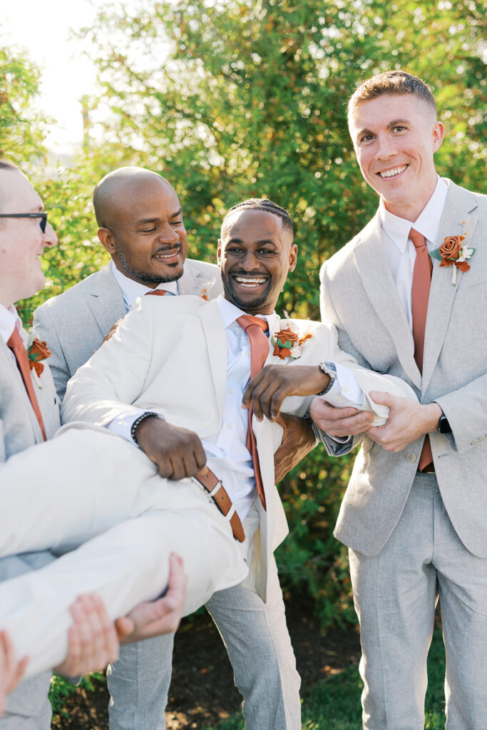 Group of three groomsmen in light gray suits and rust colored ties hold up a groom wearing a cream colored suit
