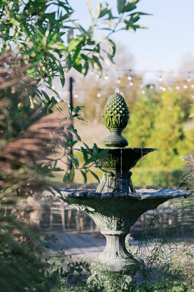 Stone fountain with a pineapple carving design on the top sits among greenery with wedding ceremony chairs and market lights in the background