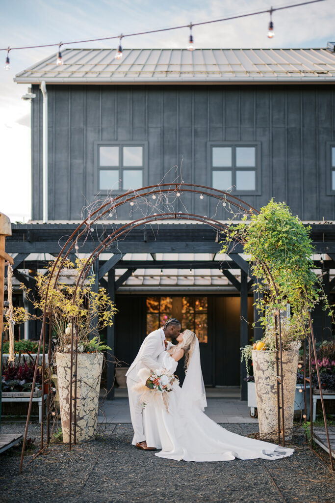 Groom dips bride and kisses her under an iron archway strung with market lights with Terrain Gardens at DelVal wedding venue in the background