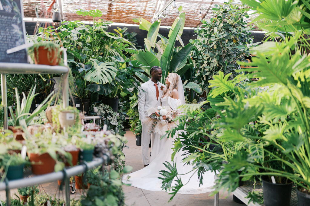 Bride and groom stand in the greenhouse at Terrain Gardens at DelVal surrounded by tall plants and greenery on all sides
