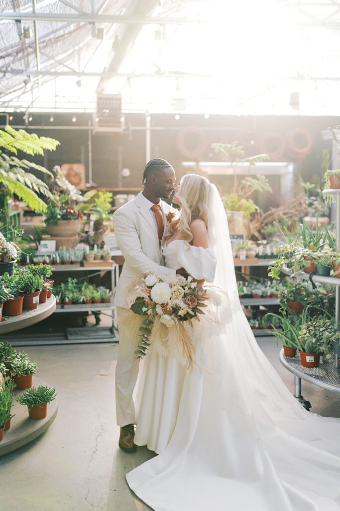 Bride and groom stand in the middle of a greenhouse surrounded by potted plants and greenery with the setting sun casting a warm hazy glow around them at Terrain Gardens at DelVal