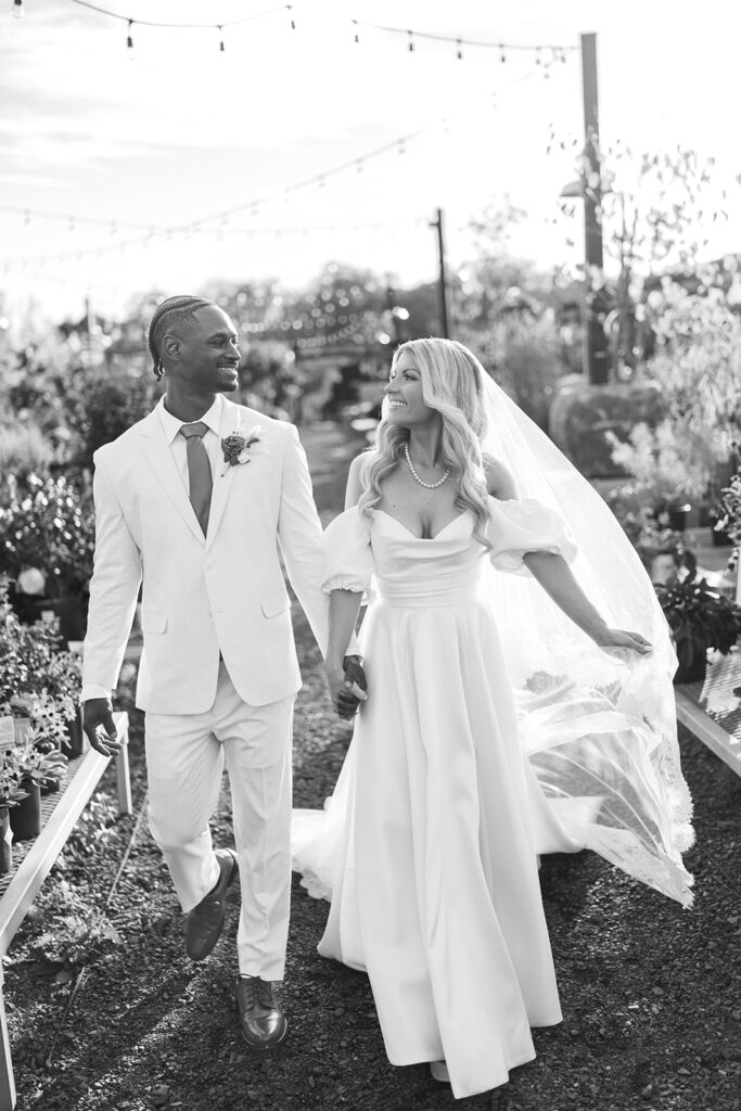 Bride and groom hold hands and smile at each other as they walk down a gravel path with market lights strung overhead at Terrain Gardens at DelVal