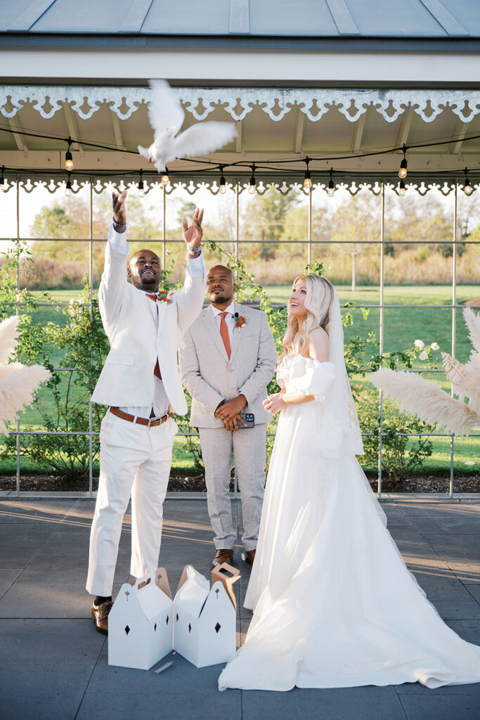 Groom releases two white doves from white boxes while his bride looks on in amazement during their wedding ceremony at Terrain Gardens at DelVal