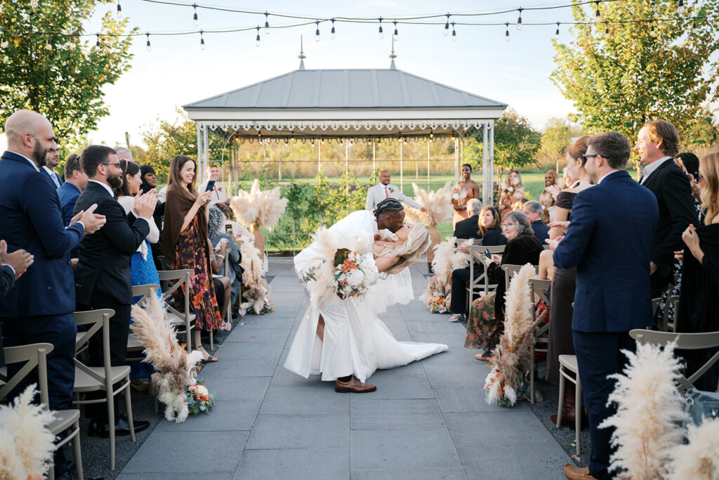 Groom dips bride and kisses her passionately in the ceremony aisle as guest smile and clap at Terrain Gardens at DelVal