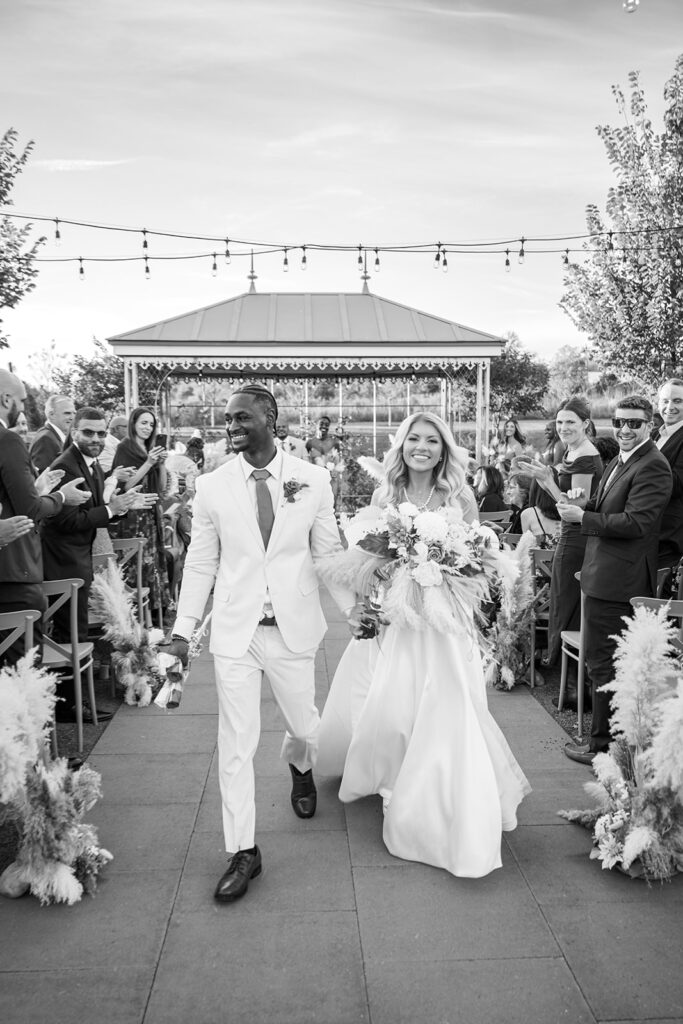 Black and white portrait of bride and groom walking down their ceremony aisle and smiling at all of their guests looking on at Terrain Gardens at DelVal