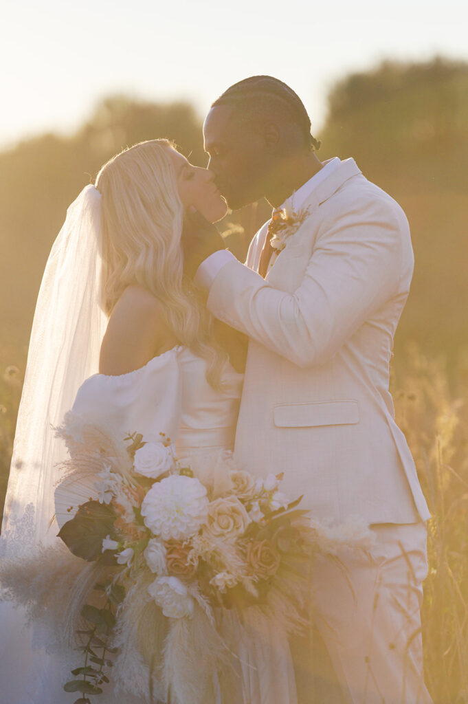 Bride and groom kiss in the middle of a field of tall grasses with the setting sun casting a warm hazy glow around them