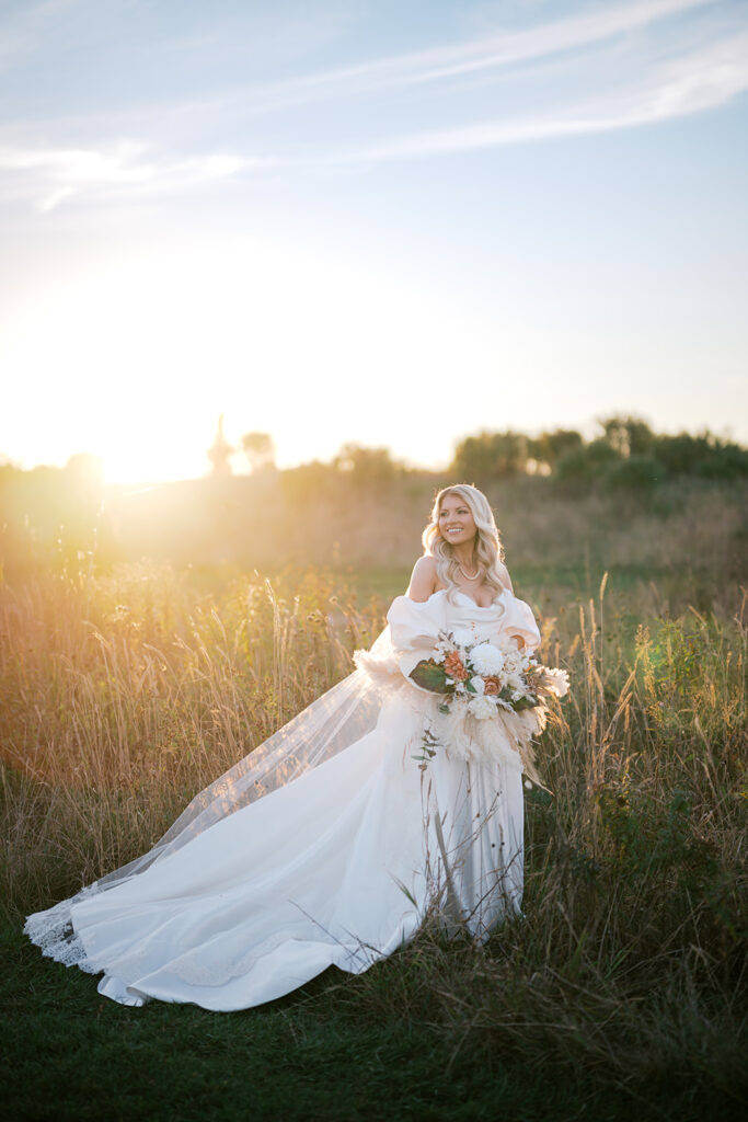 Bride poses holding a large fall floral bouquet in the middle of a grassy field while the sun sets in the background
