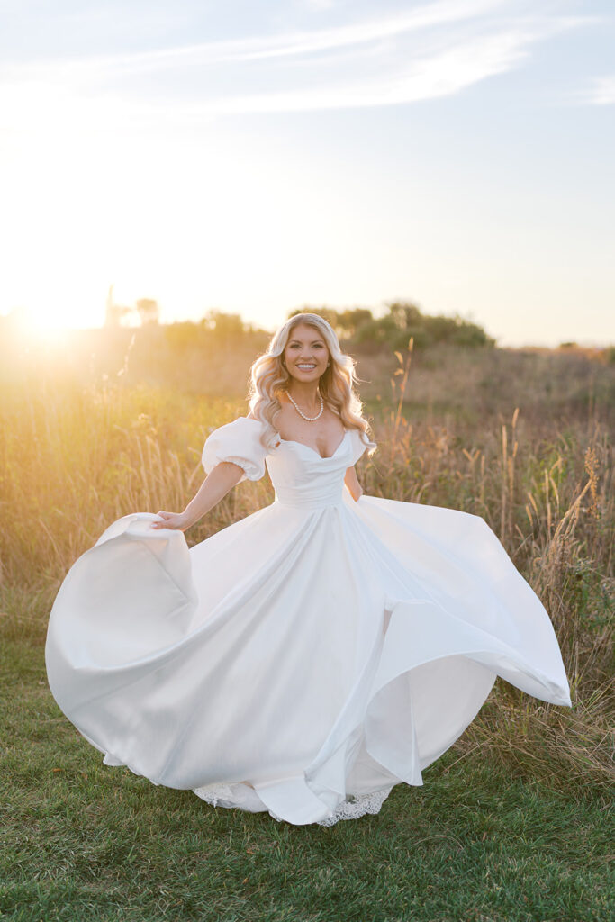 Bride twirls in a satin off the shoulder white wedding dress in the middle of a grassy field at Terrain Gardens while the sun sets in the background