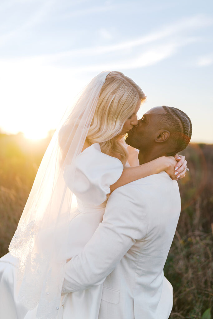 Groom holds bride up as they kiss in the middle of a field of tall grasses with the setting sun casting a warm hazy glow around them