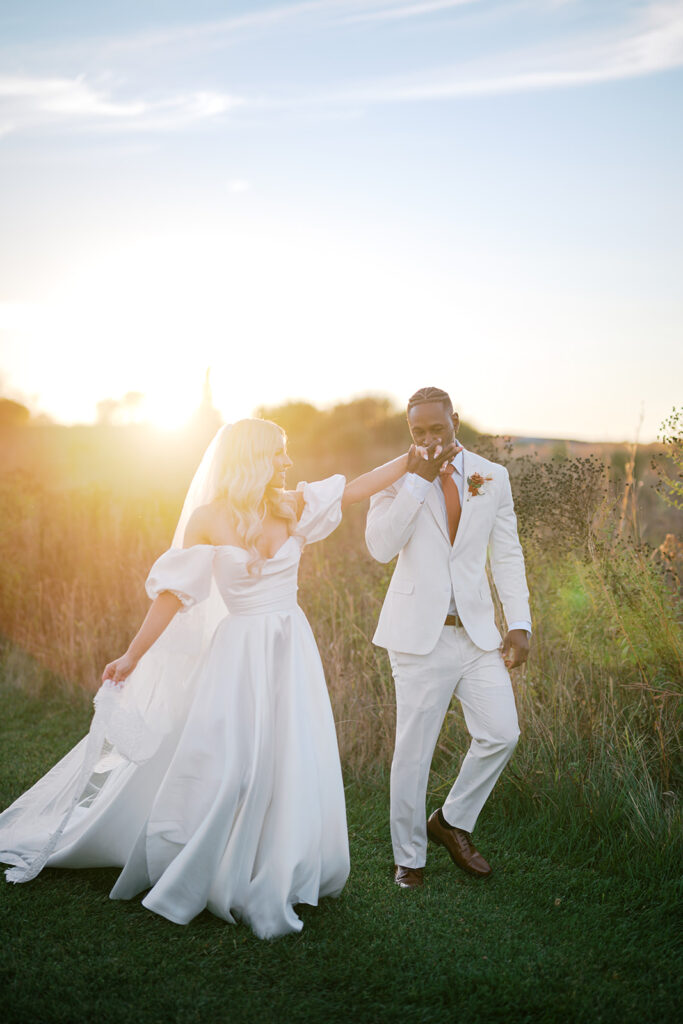 Groom kisses brides hand as they walk through a field of tall grasses with the setting sun casting a warm hazy glow around them at Terrain Gardens at DelVal