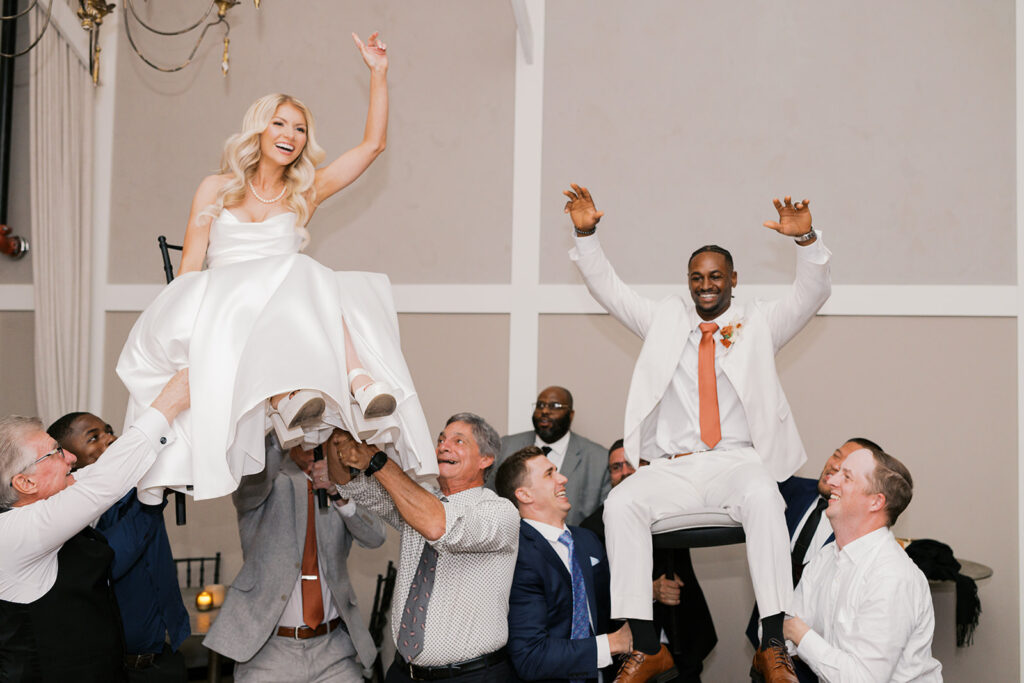 Bride and groom lift their arms and laugh as they are lifted up in a Hora style chair lift during their wedding reception at Terrain Gardens at DelVal