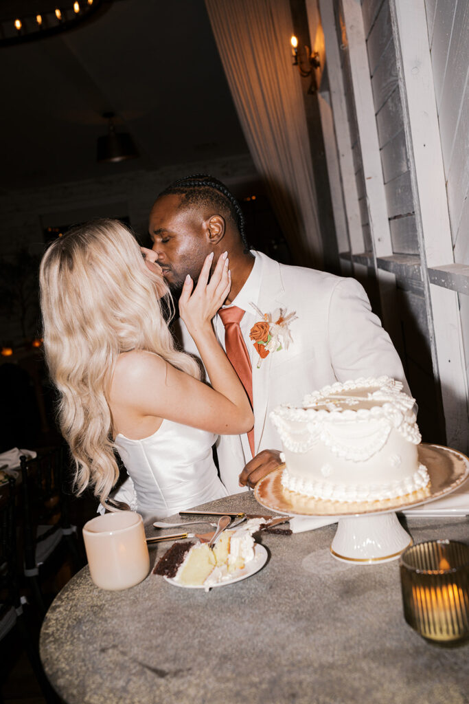 Bride holds grooms face as they kiss behind a white cake stand holding a vintage inspired buttercream piped heart shaped wedding cake